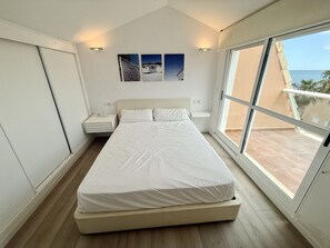 A bedroom with a large bed, a nightstand, a wall-mounted shelf, and a window with a view of the sea.