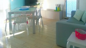 Una sala de estar con un sofá, una mesa de comedor con sillas, un televisor y un cuadro que representa una playa.