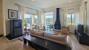 Een ruime woonkamer met een bank, stoelen, een televisie en een open haard. Grote ramen bieden uitzicht op zee en heuvels.