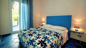 A hotel room with a bed, a nightstand with a lamp, a window with curtains, and a view of the outdoors. - Seaview 3-room apartment- 3 min by car from the beach (Capo d'Orlando)