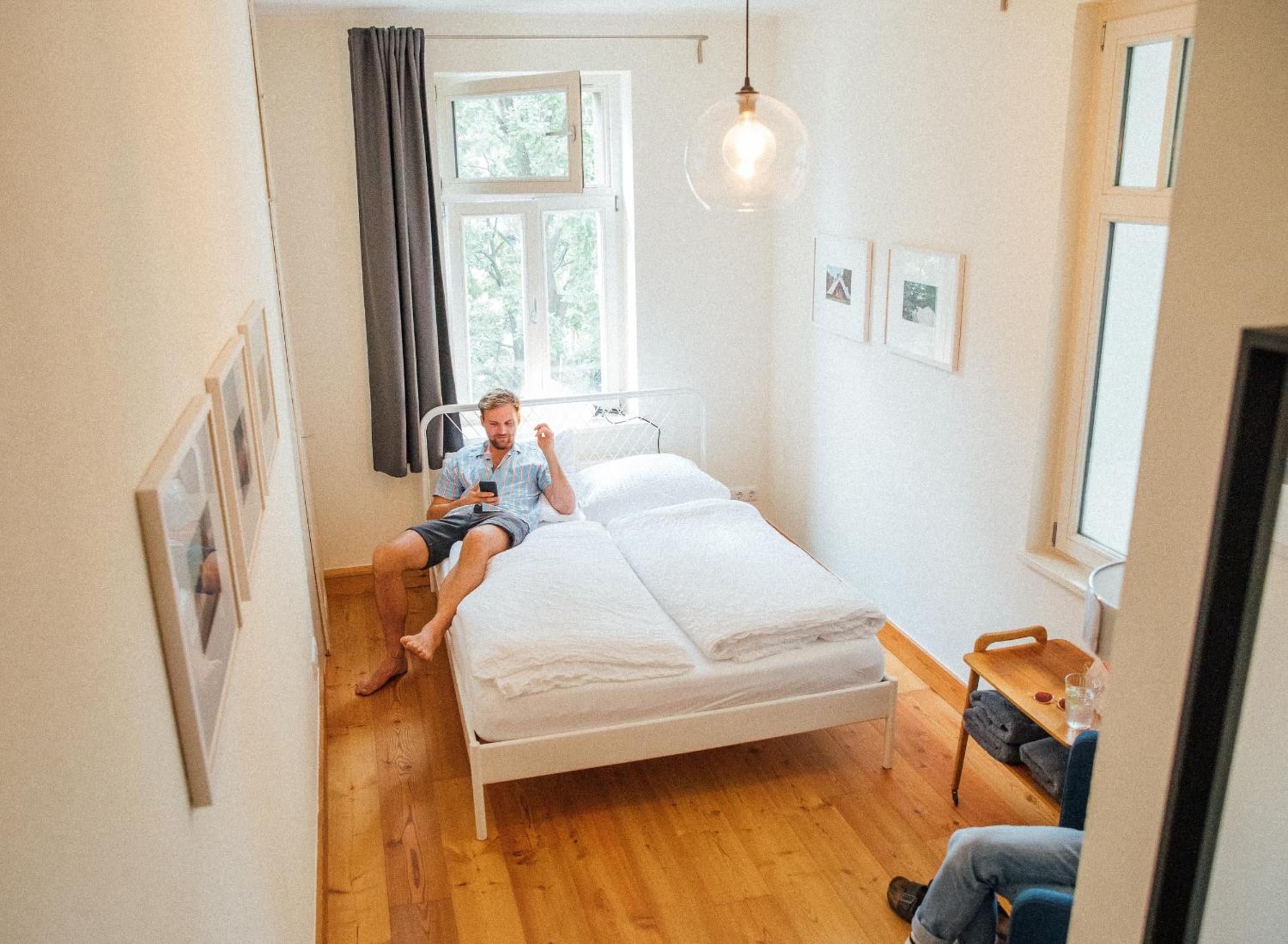 A single-bed room with a wooden floor, a bedside table with a glass, and a hanging light fixture.