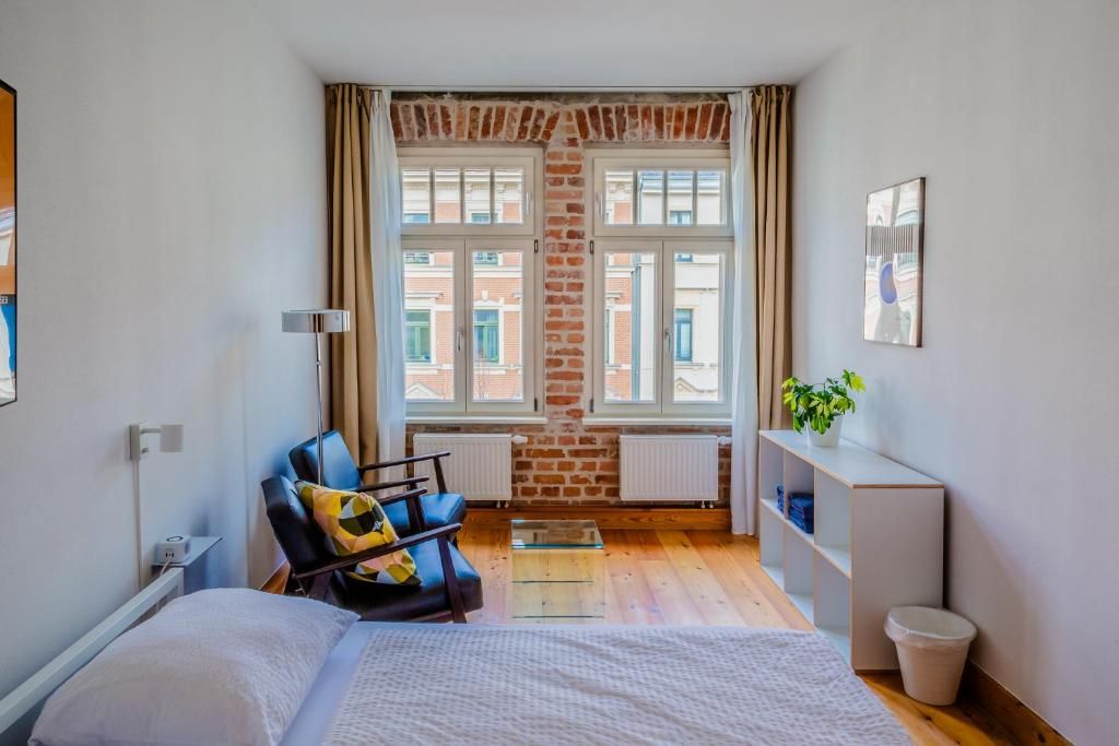Een compacte hotelkamer met een bed, een zithoek met een stoel en een tafeltje, een radiator, een raam met gordijnen en een wandplank met een plant.