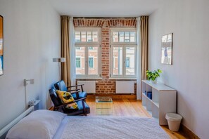 A compact hotel room with a bed, a seating area with a chair and a small table, a radiator, a window with curtains, and a wall-mounted shelf with a plant. - Hostel Multitude (Leipzig)