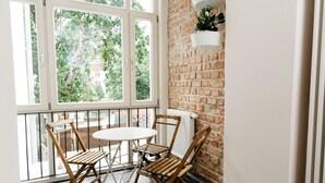 A balcony with two wooden chairs and a small round table, a brick wall, and a window overlooking greenery.