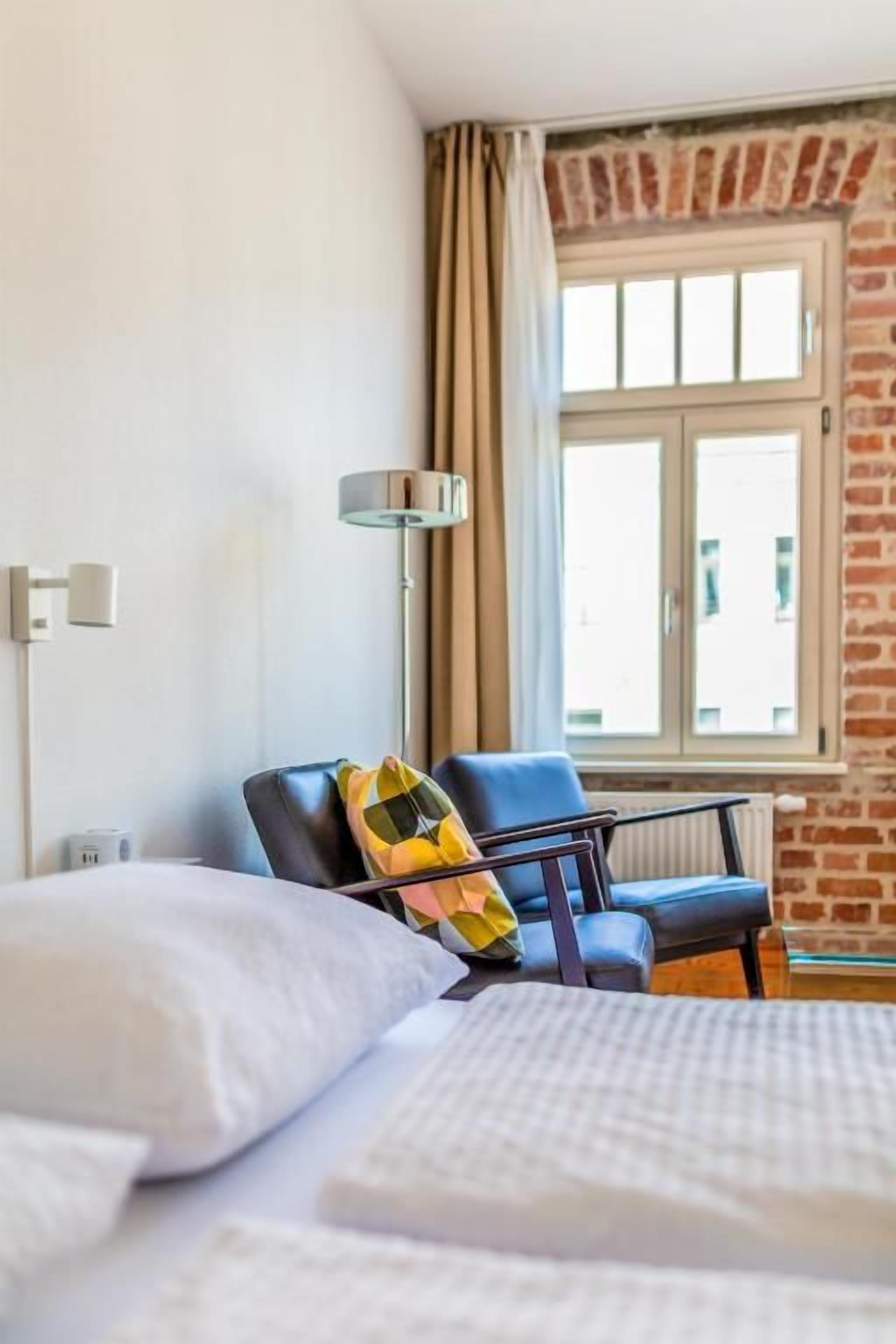 A hotel room with a bed, a chair, a window with curtains, and a lamp.