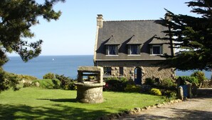 Ein Steinhaus mit schrägem Dach, einem Stebrunnen und Blick auf das Meer.