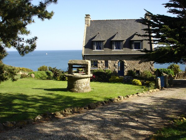 A stone building with a sloped roof, a stone well, and a view of the sea.