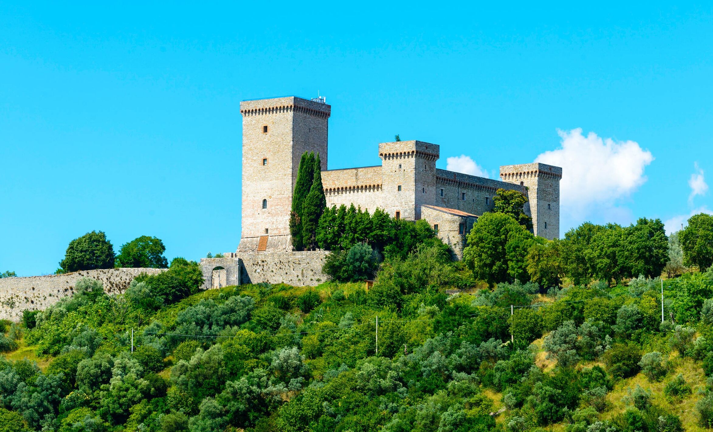 Un château de pierre historique, doté de nombreuses tours, entouré d’une végétation luxuriante et surplombé d’un ciel bleu azur.
