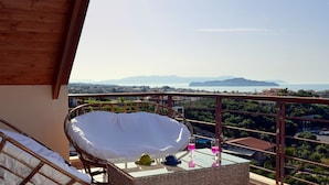 Un balcone con una sedia in vimini, un tavolino in vetro e vista su una cittadina costiera e montagne.