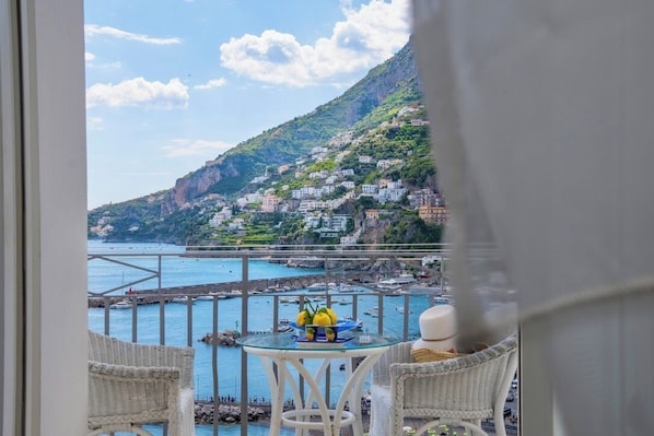 A balcony with wicker chairs, a small table with fruit, and a view of a coastal town with buildings on a hillside.