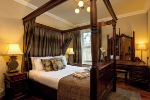 A four-poster bed with a canopy, a wooden desk with a mirror, and a bedside table with a lamp.