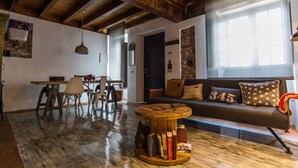 Ein gemütlicher Wohnbereich mit Holzboden, einem runden Couchtisch mit Büchern, einem Sofa mit Kissen in Sternenmuster und einem Essbereich mit Stühlen und einem hängenden Blumentopf.
