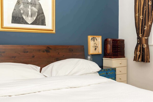 Een net opgemaakt bed met witte lakens, een houten hoofdbord, een ingelijst foto op de muur, een nachtkastje met een lamp, en een blauwe decoratieve doos.