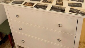 Eine weiße Kommode mit Glasplatte, auf der Fotografien ausliegen, flankiert von zwei hölzern gerahmten Spiegeln an der Wand.
