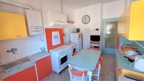 A compact kitchen with a sink, stove, microwave, and a small refrigerator. There is a dining table with chairs and a clock on the wall.