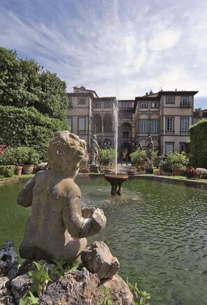 Eine Steinstatue eines Löwen in einem Brunnen, umgeben von Grünpflanzen und einem Gebäude mit klassischer Architektur im Hintergrund.