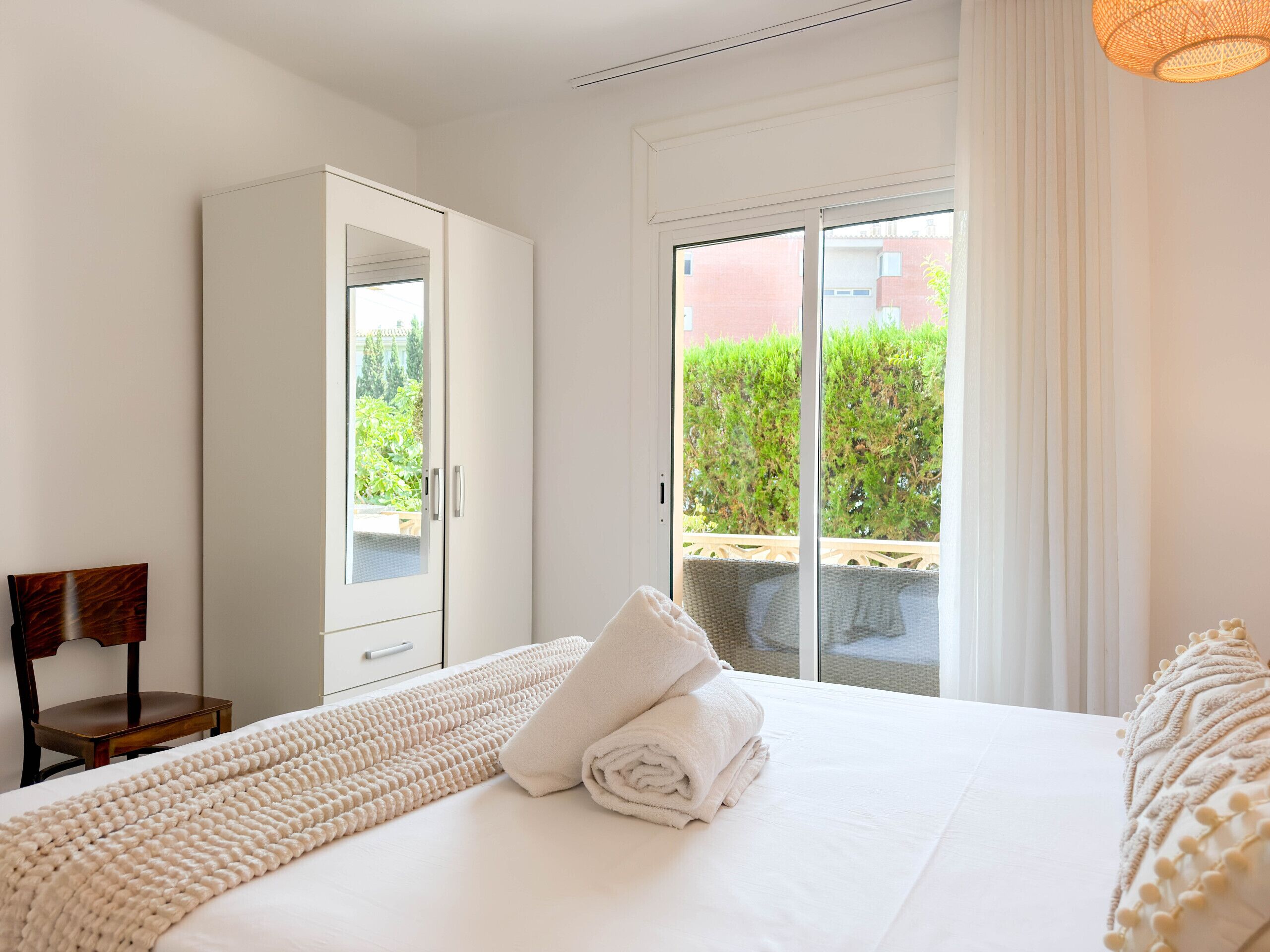 Ein Schlafzimmer mit Bett, Kleiderschrank, Stuhl und Blick ins Grüne durch eine Schiebetür.