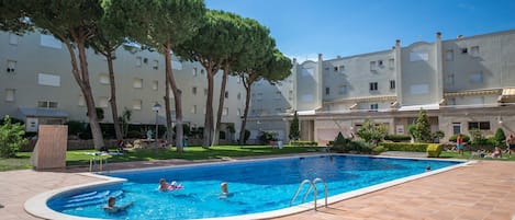 A swimming pool surrounded by a courtyard with trees and a building complex in the background.