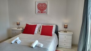 Ein ordentlich bezogenes Bett mit gestreifter Bettwäsche und roten Kissen, zwei Nachttische mit Lampen, ein weißer Beistelltisch und ein gerahmtes Kunstwerk an der Wand.