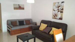 Ein Wohnzimmer mit grauem Sofa, einem dunklen Sofa und einem kleinen Tisch mit Gläsern darauf. Auf den Sofas liegen Kissen und an der Wand hängt ein Gemälde.