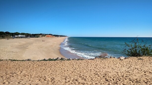 Ein weiter Sandstrand mit sanften Wellen, blauem Himmel und einigen Spaziergängern.