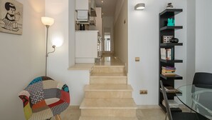 Un intérieur moderne avec un escalier menant à un espace cuisine, une table en verre et une bibliothèque contenant des livres et des objets décoratifs.