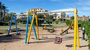 A playground with swings and a slide, set against a backdrop of palm trees and a building. - Lovely appartment at 50m from the beach, with swimming pool and beach bar. 4 pers. Wifi. (Dénia)