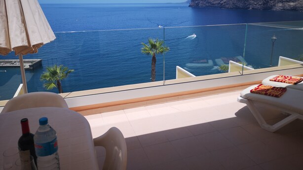 Ein Balkon mit freiem Blick auf das Meer und ein Berg. Ein Tisch mit einer Flasche und einem Glas sowie ein Stuhl sind vorhanden.