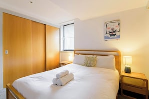 A neatly made bed with rolled towels, a wooden headboard, a bedside lamp, and a window with blinds. - Modern 2BDR Flat by Cathedral in Old Town (Edinburgh)