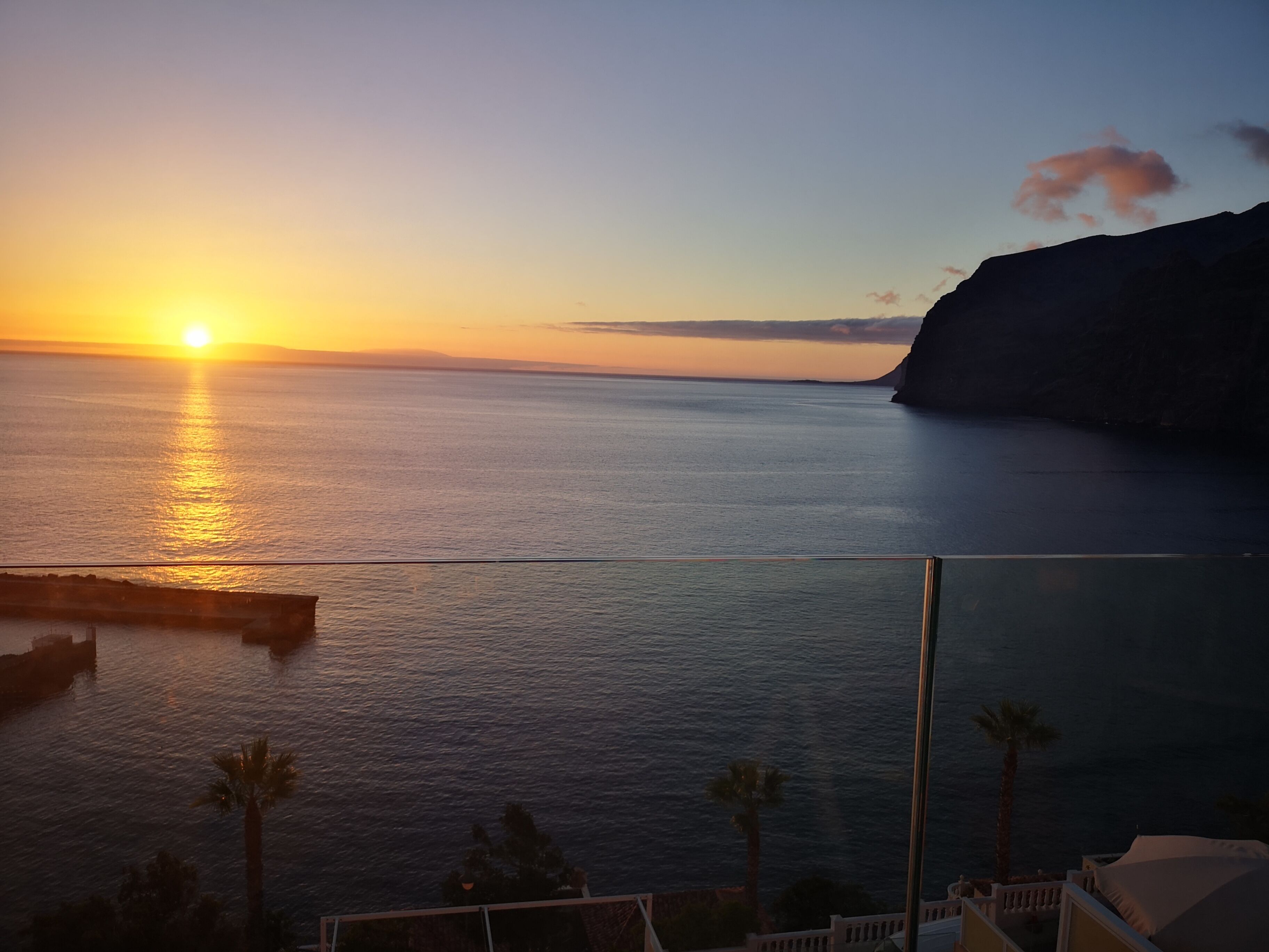 Vista do pôr do sol a partir de uma varanda com vista para um mar calmo, um penhasco ao longe e palmeiras.