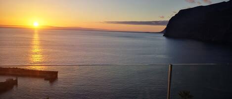Vista do pôr do sol a partir de uma varanda com vista para um mar calmo, um penhasco ao longe e palmeiras.