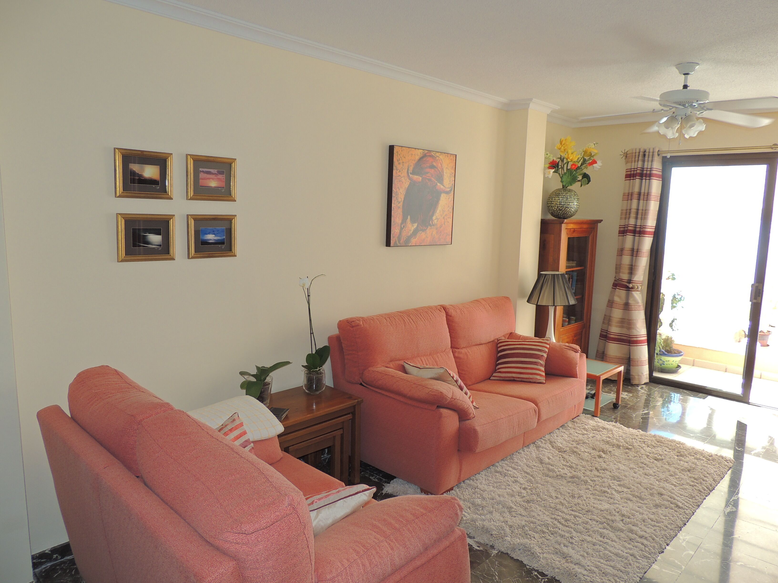 A living room with a sofa, armchairs, a coffee table, and a TV. There are framed pictures on the wall, a ceiling fan, and a window with curtains.
