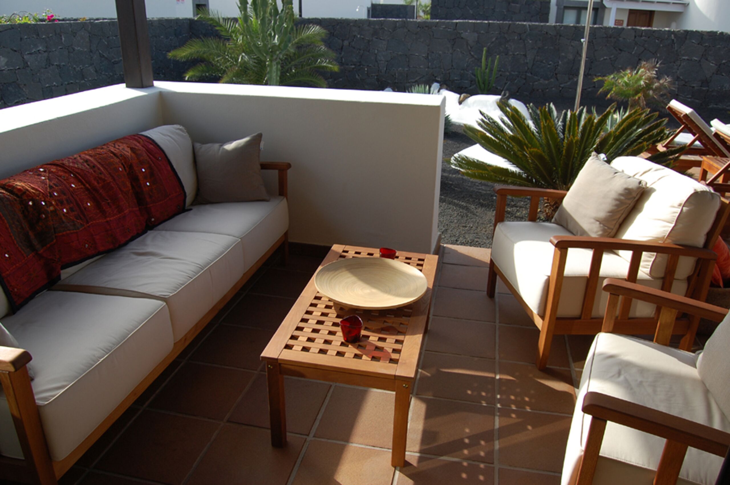 A sunlit patio with a sofa, two wooden chairs, and a table with a bowl and a red cup.