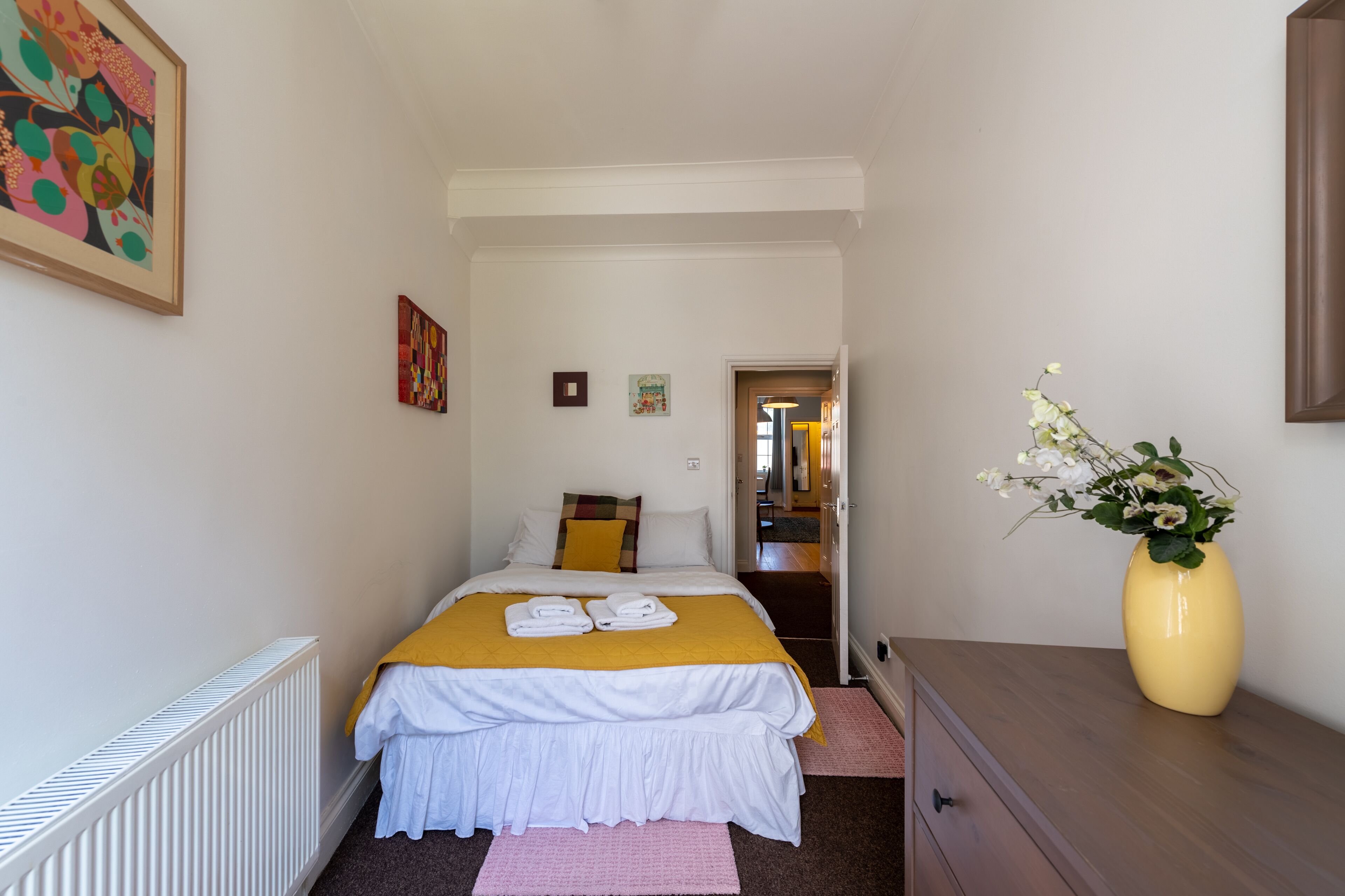 A bedroom with a bed, a dresser, a radiator, a vase with flowers, and a hallway leading to another room.
