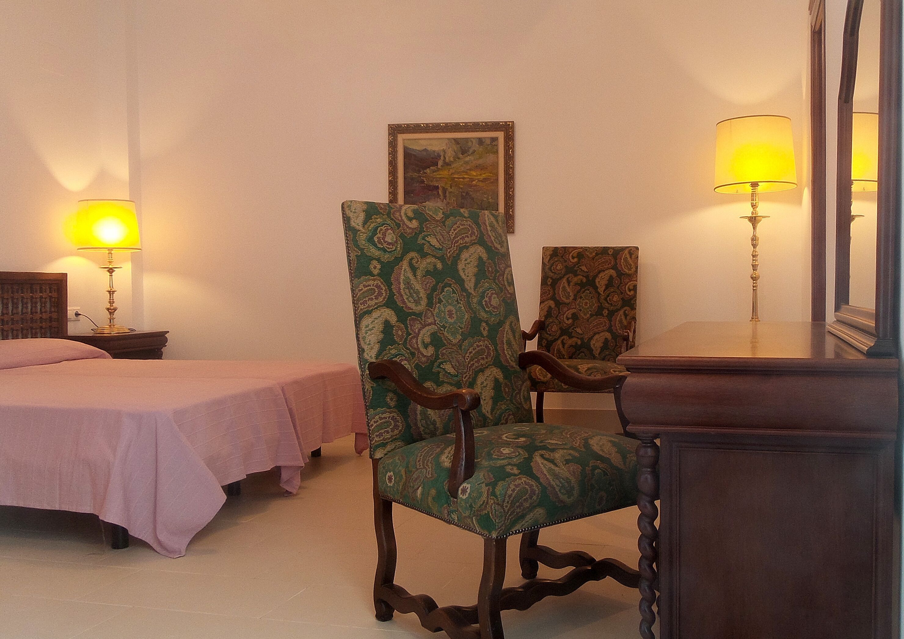 A hotel room with two single beds, a green patterned armchair, a wooden nightstand with a lamp, and a mirror.
