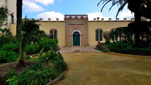 Un bâtiment doté d’une entrée centrale, entouré d’un jardin où poussent différentes espèces de plantes, et un chemin pavé menant à l’entrée.
