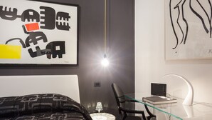 Une chambre d’hôtel moderne, équipée d’un lit, d’un bureau avec un ordinateur portable, d’une chaise, d’une lampe et ornée d’œuvres d’art abstraites sur les murs.