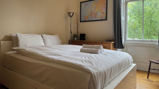 A bedroom with a large bed, a nightstand, a lamp, a map on the wall, and a window with a view of trees. - Charming Morningside Flat in Edinburgh (Edinburgh)
