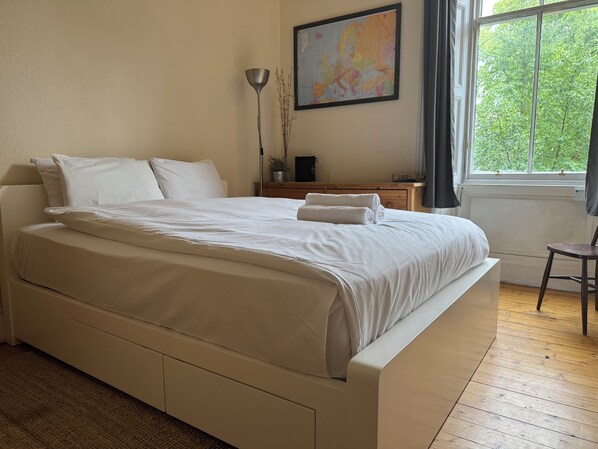 Cama bem arrumada em quarto com piso de madeira, mesa de cabeceira com luminária e janela com vista para árvores.