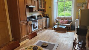A kitchen with wooden cabinets, a dining table with a floral placemat, and a view of greenery through a window. - Charming Morningside Flat in Edinburgh (Edinburgh)