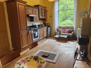 A kitchen with wooden cabinets, a dining table with a floral placemat, and a view of greenery through a window. - Charming Morningside Flat in Edinburgh (Edinburgh)