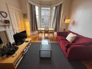 A living room with a TV, red sofa, glass coffee table, wooden TV stand, fireplace, dining table, and curtains.