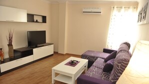 Una sala de estar con televisor de pantalla plana, una mesa de centro blanca, un sofá y una mesita con un florero y control remoto.