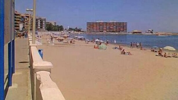 Una playa con un edificio grande al fondo, cielo despejado y gente relajándose en la arena.
