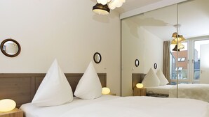 Habitación de hotel con un espejo grande, dos mesitas de noche con lámparas, una cama con ropa de cama blanca y vistas al exterior a través de una ventana.