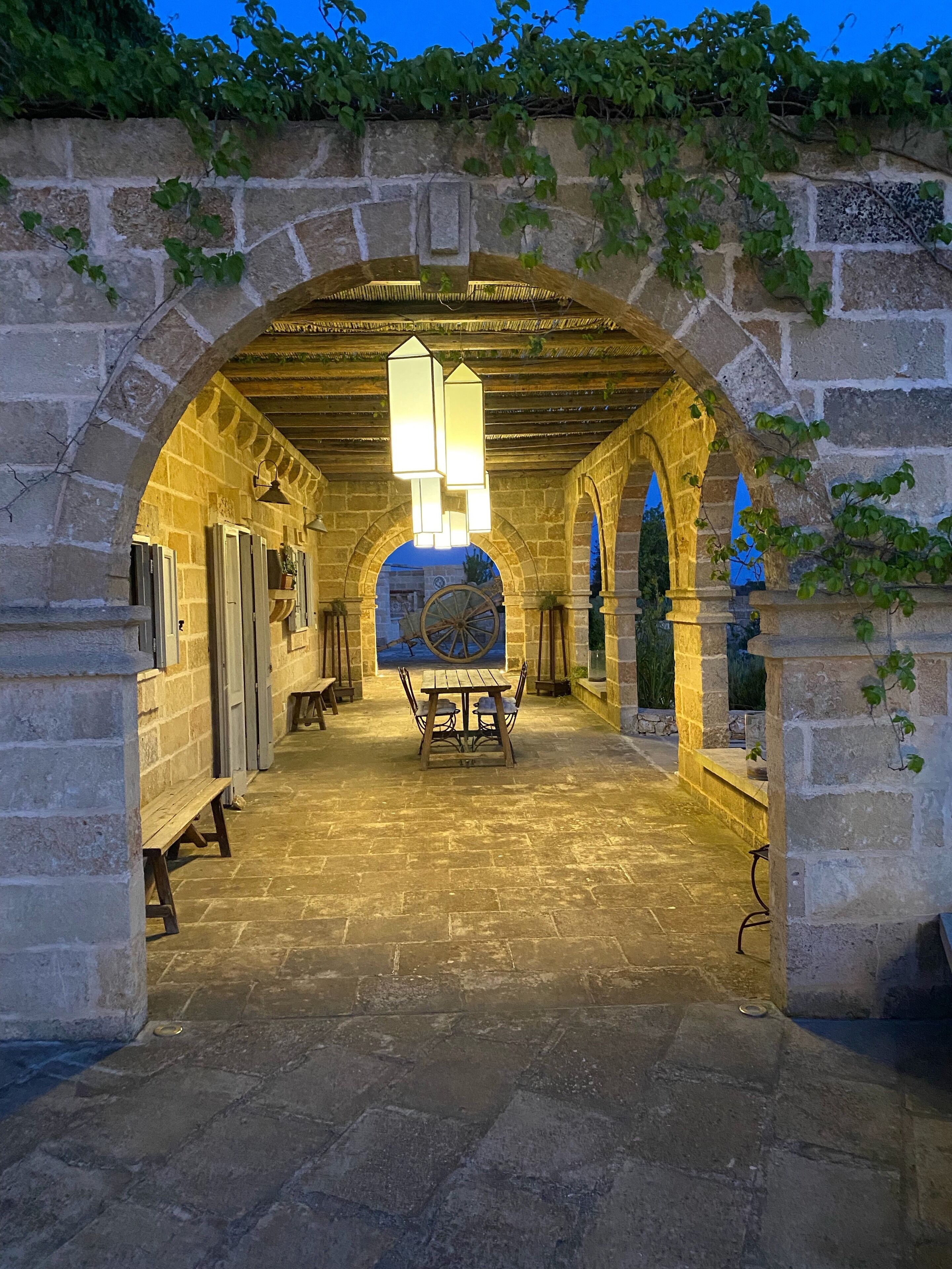 Un corridoio in pietra con archi, un tavolo in legno, panche e lanterne sospese.