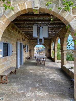 Un porche techado al aire libre con paredes de piedra, una mesa de madera con sillas y una rueda grande al fondo.