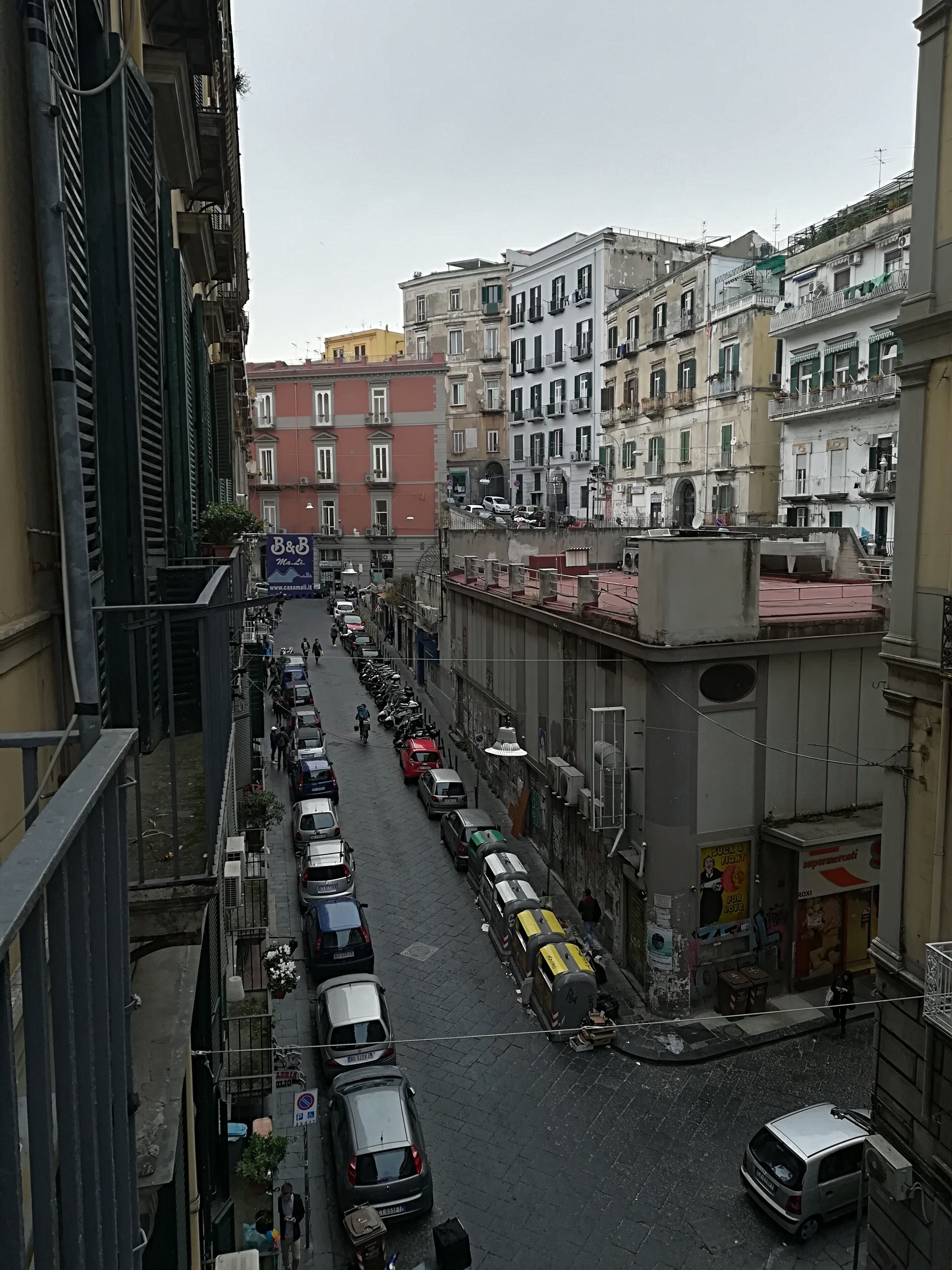A narrow urban street lined with multi-story buildings, parked cars, and a few pedestrians.