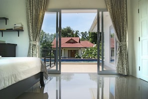Un dormitorio con cama, vista a través de un ventanal hacia un área de piscina y un edificio de techo rojo al fondo.