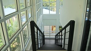 Una escalera con barandillas negras que sube a un segundo piso, rodeada de grandes ventanales con vistas a zonas verdes y edificios.
